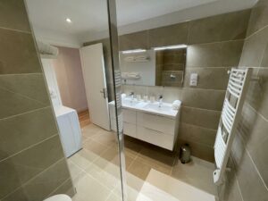 Salle de bain rénovée avec douche à l'italienne, double vasque, carrelage beige et espace buanderie.