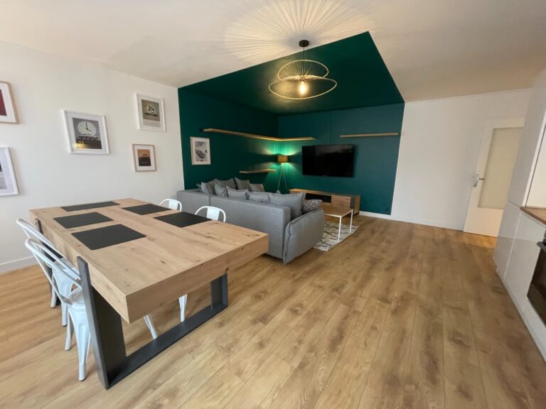 Salon et salle à manger rénovés avec mur vert, table en bois, canapé gris et télévision murale.