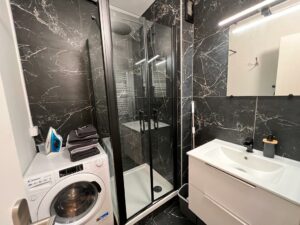 Salle de bain rénovée avec douche moderne, carrelage noir marbré, machine à laver et lavabo blanc.